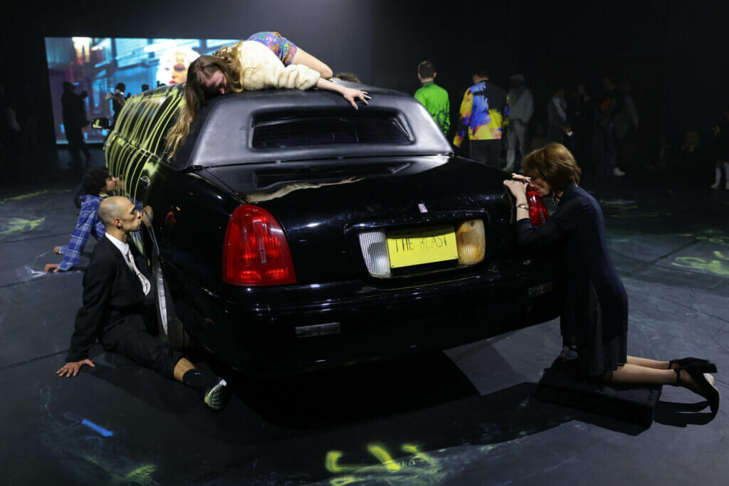 Tänzer:innen knien um ein Auto herum. Das Auto ist mit Graffiti besprüht. Auch auf dem Boden sind gesprühte Zeichen zu sehen.