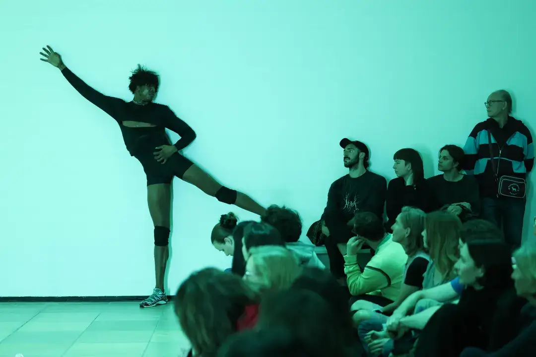 Eine Person hebt an der linken Seite der Bühne das Bein zur Arabesque. Rechts sitzt eine Gruppe von Menschen, die dabei zuschauen.