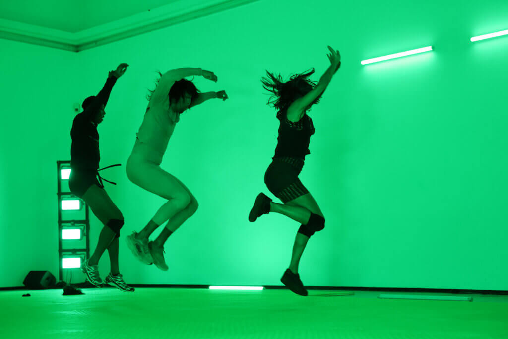 Drei Tänzer:innen hübfen hintereinander her. Ihre Arme sind jubelnd über die Köpfe gehoben.