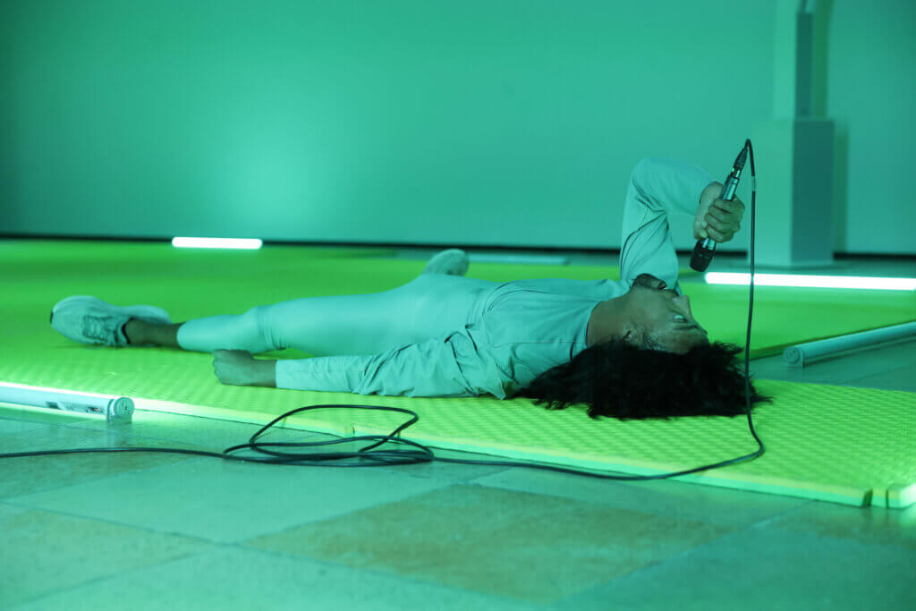 Ein Tänzer liegt auf dem Rücken, in der Hand ein Mikrofon, in das er spricht.