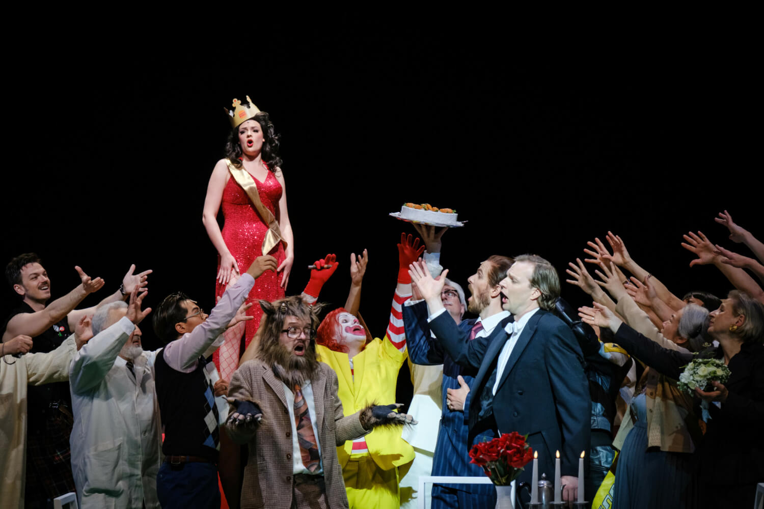 Eine Frau steht auf einer Erhebung im langen Abendkleid mit Scherpe und Krone auf der Bühne. Um sie herum ist eine diskutierende Menge versammelt, unter anderem sind ein Clown, ein Wolfskopf und Anzugträger zu sehen.