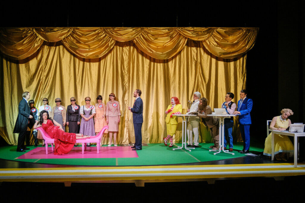 Schönheitskönigin Diana Deveraux liegt auf der linken Bühnenseite auf einer Liege. Hinter ihr sind weitere Kandidatinnen aufgereiht. Rechts steht eine Jury am Tisch.