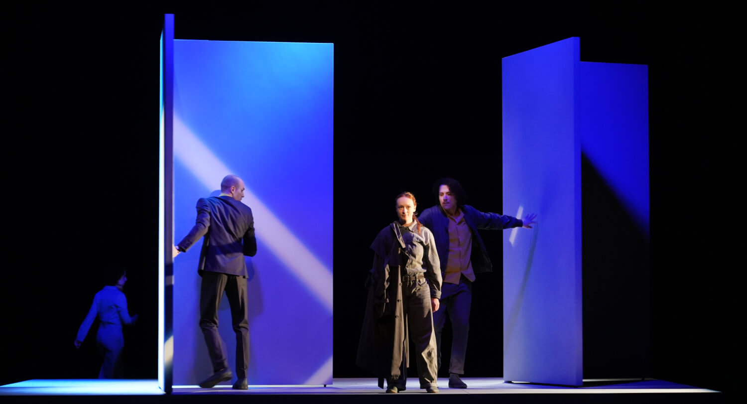 Die Bühne ist dunkel ausgeleuchtet. In der Mitte steht eine Frau in Geheimdienstkleidung und schaut ins Publikum.