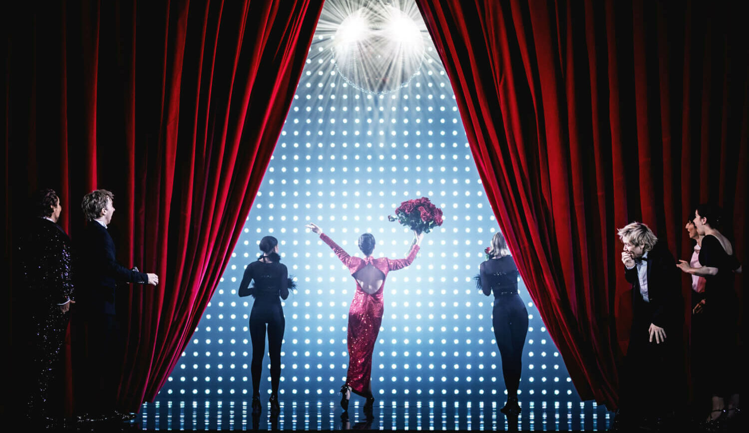 An der Bühnendecke hängt eine schillernde Diskokugel. Rechts und links hängt ein Bühnenvorhang. In der Mitte der Bühne steht eine Darstellerin mit einem Strauß Rosen, den sie hoch hält.