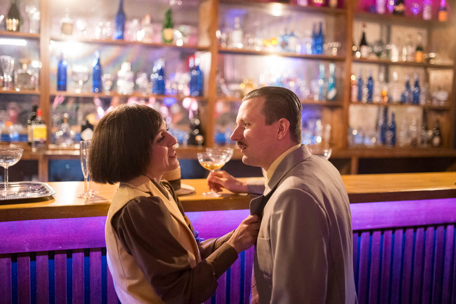 Im Hintergrund ist eine Bar mit allerlei Flaschen und Gläsern beleuchtet. Vorne stehen eine Frau und ein Mann und schauen sich an.