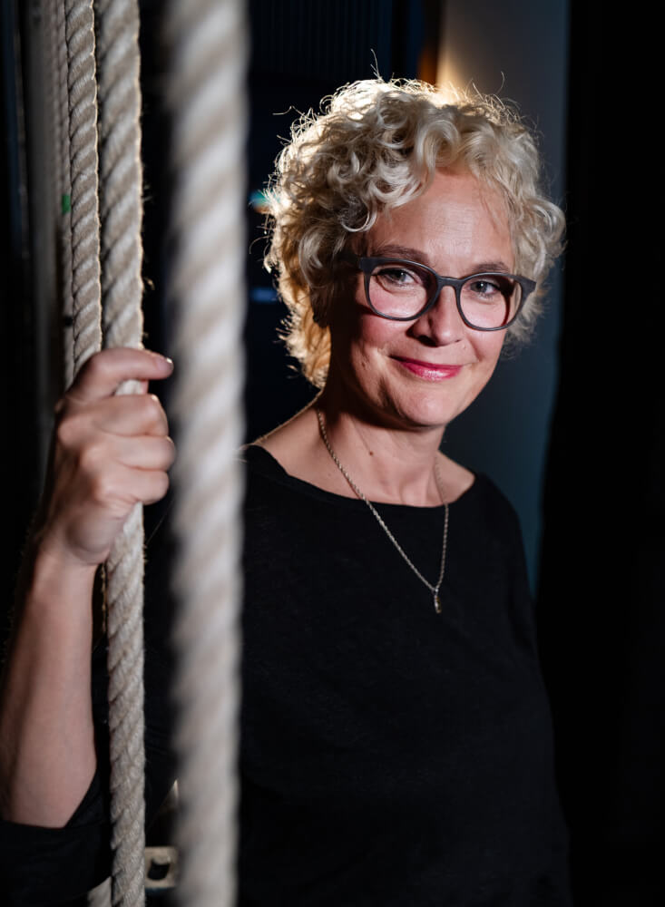 Zu sehen ist Sarah Kohrs, Intendantin des Landestheater Schwaben. Mit der rechten Hand hält sie sich an einem von mehreren Seilzügen fest. Sie trägt eine schwarze Brille und ein schwarzes Oberteil.