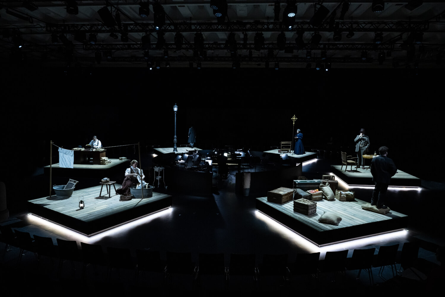 Szenenfoto aus „The Strangers“ an der Oper Köln. Zu sehen sind sechs Podeste, die indirekt von unten beleuchtet sind. Darauf sind rechts vorne Kisten und eine Person, links vorne eine Wäscheleine, Schemel und eine sitzende Frau. Witer hinten eine Straßenlaterne und eine Kirchenbank mit goldenem Kreuz.