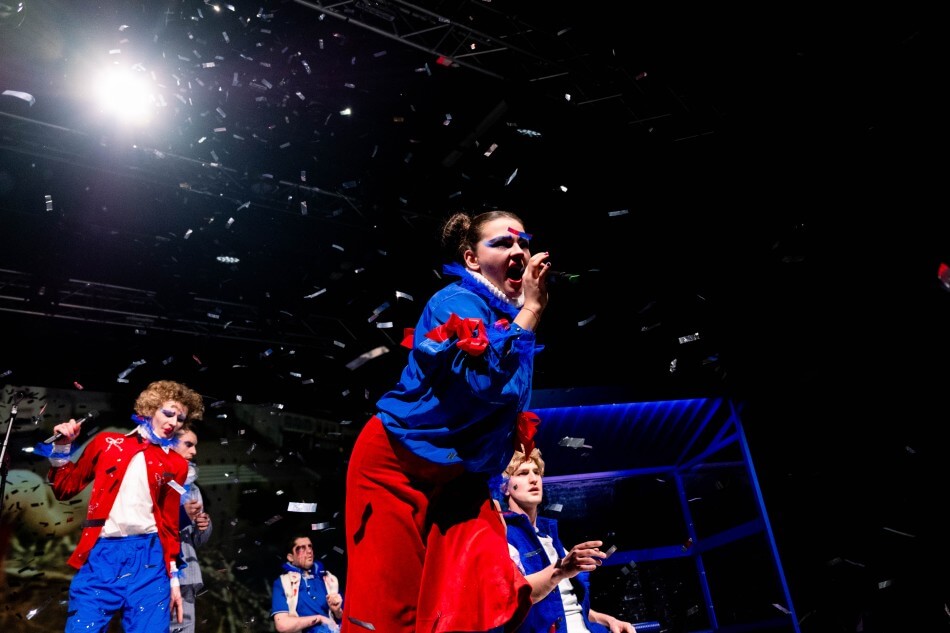 eine darstellende Person im Vordergrund, die ins Mikro spricht, dahinter eine weitere Person