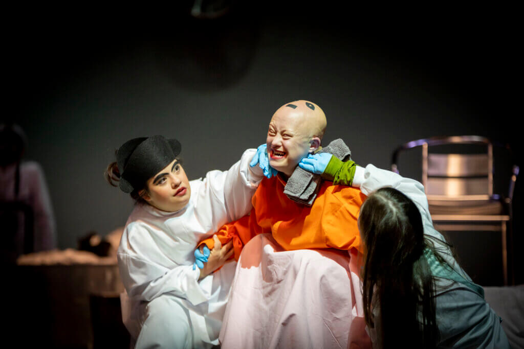 Zwei Tänzer:innen knien bei einer Person mit Glatze, die angestrengt ihr Gesicht verzerrt.