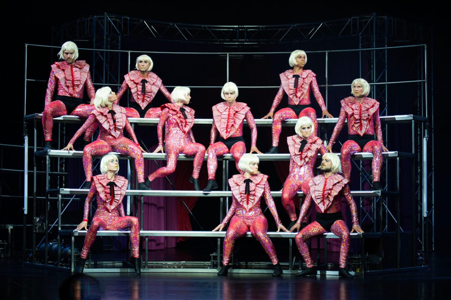 Auf einer Tribüne sitzen unterschiedliche Personen. Alle sind in pinken Anzügen und blonden Perücken gekleidet.