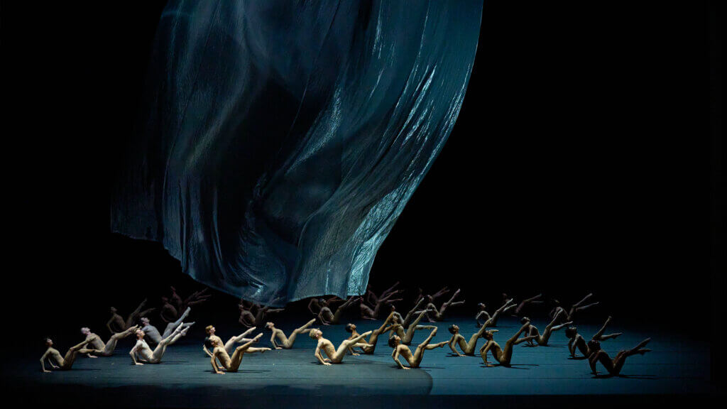 Tänzer:innen sitzen symmetrisch aufgereiht auf dem Boden und strecken die Beine wie bei einer Bauchmuskelübung von sich. Über der Gruppe schwebt eine Folie.