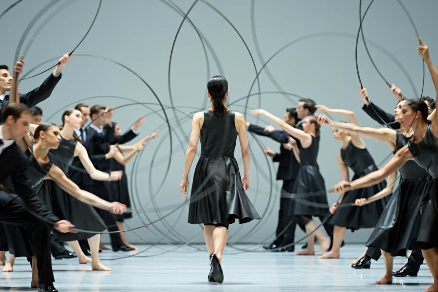 Eine Person schreitet durch eine im Spalier stehende Gruppe von der Kamera weg. Das Tanz-Ensemble wedelt hält zu Kreisen gebogene schwarze Stäbe in den Händen.