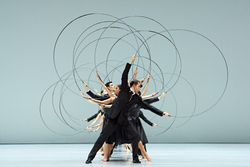 Das Ensemble steht hintereinander in einer Reihe und bewegt große Stabbögen in kreisenden Bewegungen.