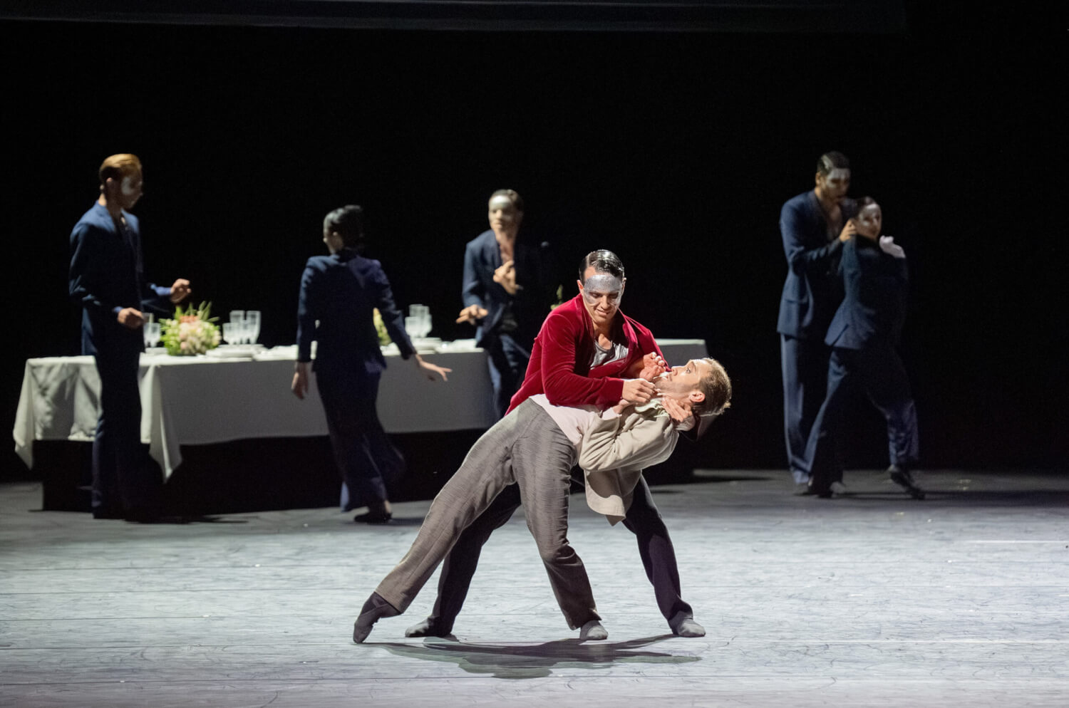 Eine Tänzerin wird von einem Tänzer zurückgebeugt. Im Hintergrund stehen Menschen in Anzügen an einem langen Tisch.