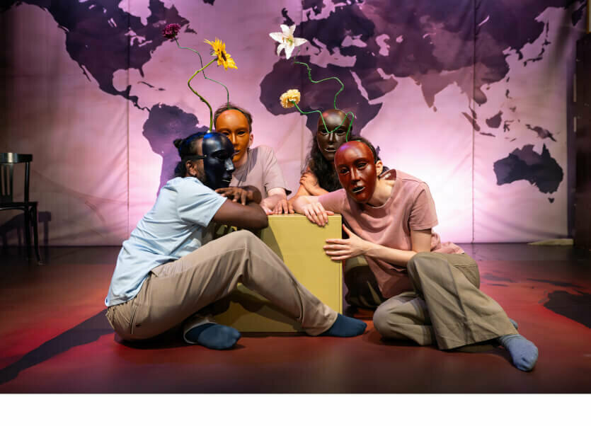 Chandrashekara Kempaiah, David Smith, Devaki Rajendran und Larissa Voulgarelis sitzen mit Holzmasken vor den Gesichtern und einer Blume aus den Köpfen ragend auf dem Boden zusammen.