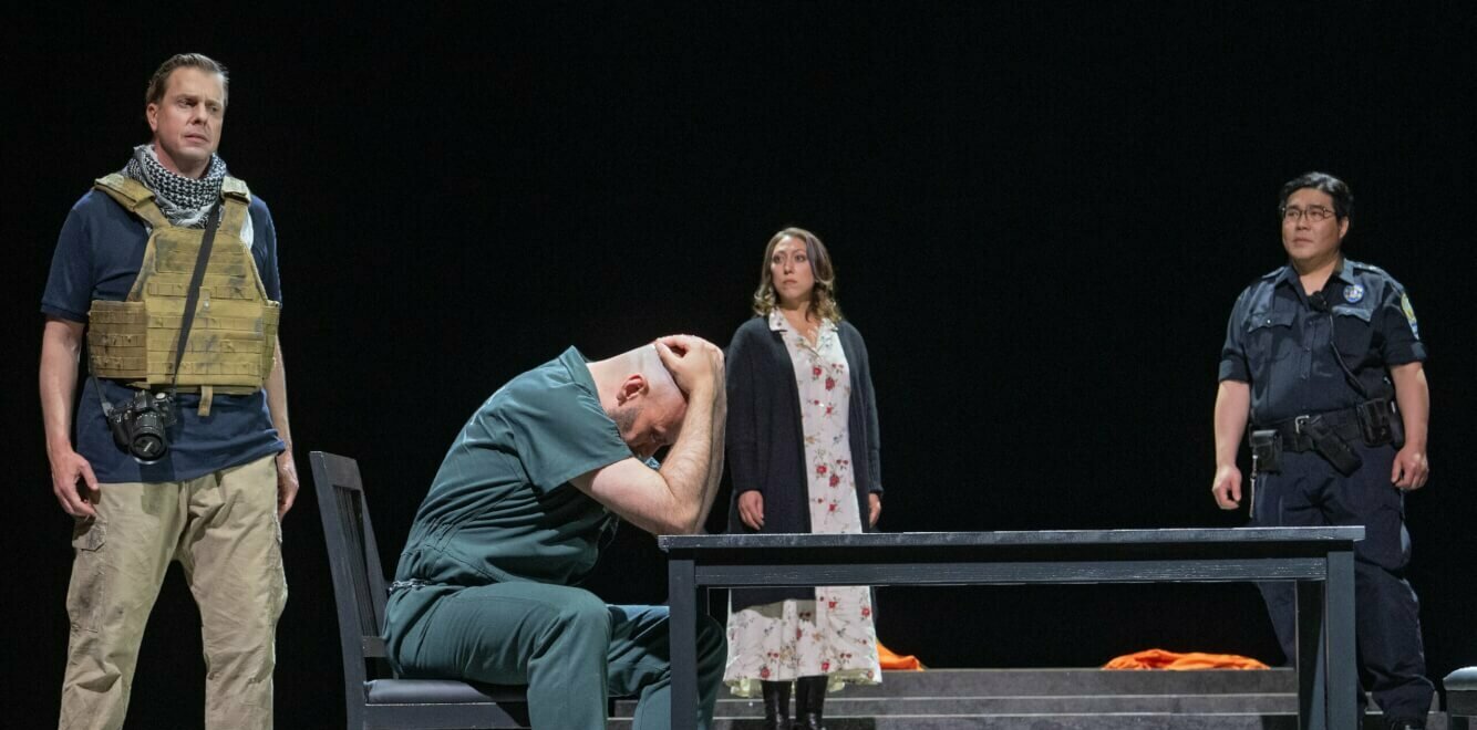 Alexanda Kotey (Timothy Connor) sitzt den Kopf in den Händen vergraben an einem Tisch. Links hinter ihm steht der tote Journalist James Wright Foley mit seiner Kamera.