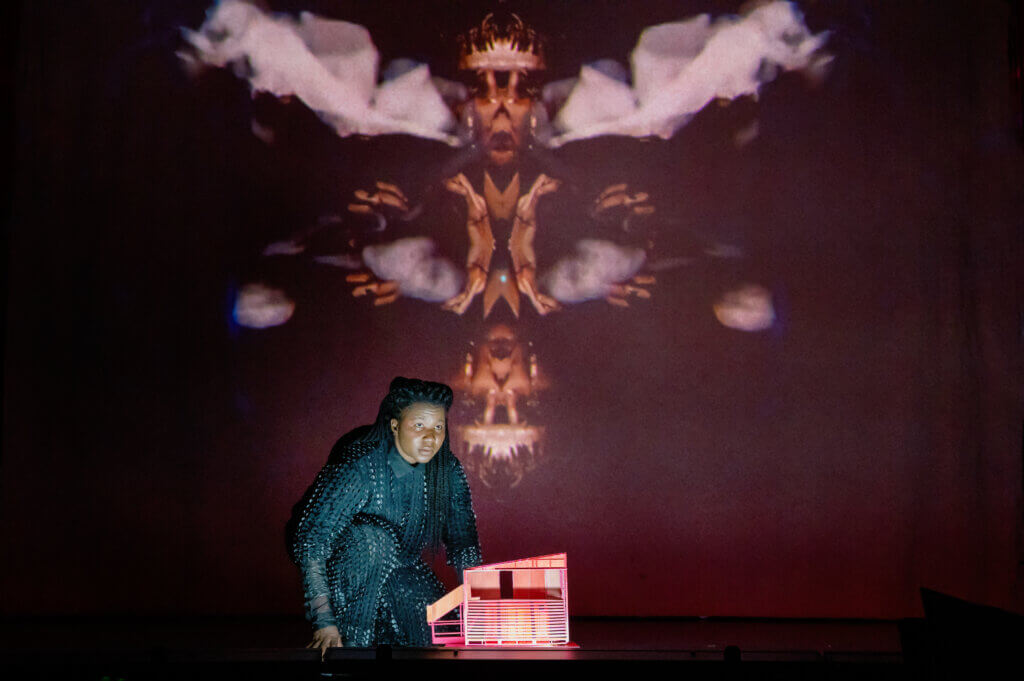 Eine Darstellerin vor einem Hintergrund. Vor ihr steht ein Puppenhaus.