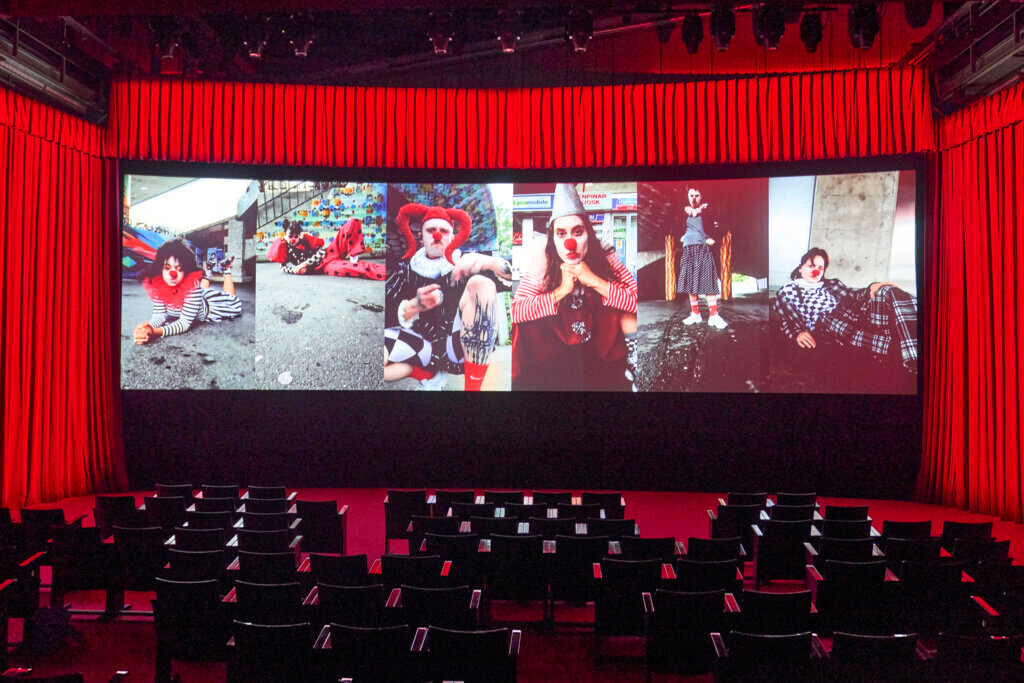 Auf einer Leinwand sind sechs Hochkant-Videoaufnahmen mit den Darstellenden zu sehen.