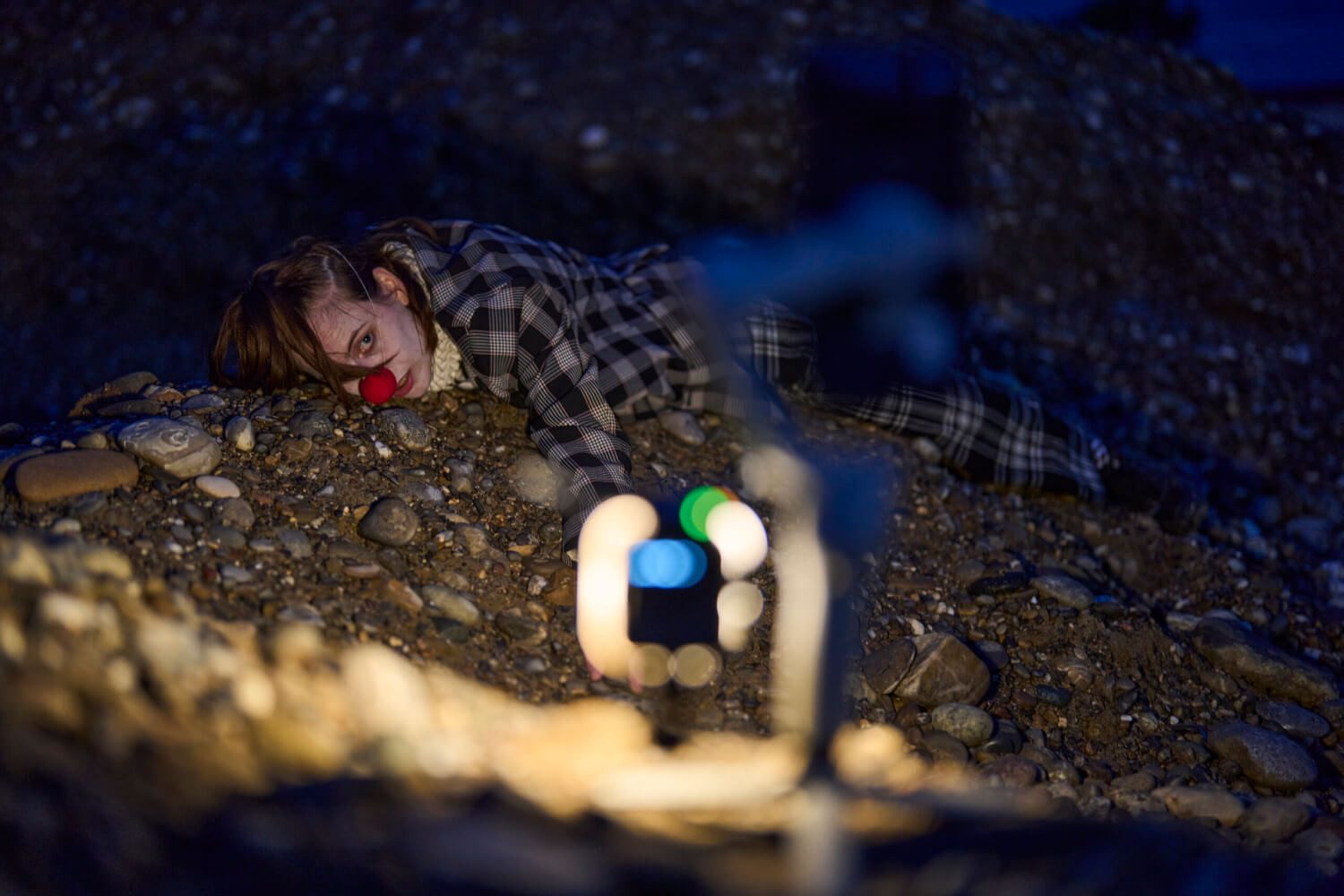 Eine junge Person liegt in ein Hemd gekleidet auf dem Asphalt. Sie trägt eine Clownsnase.