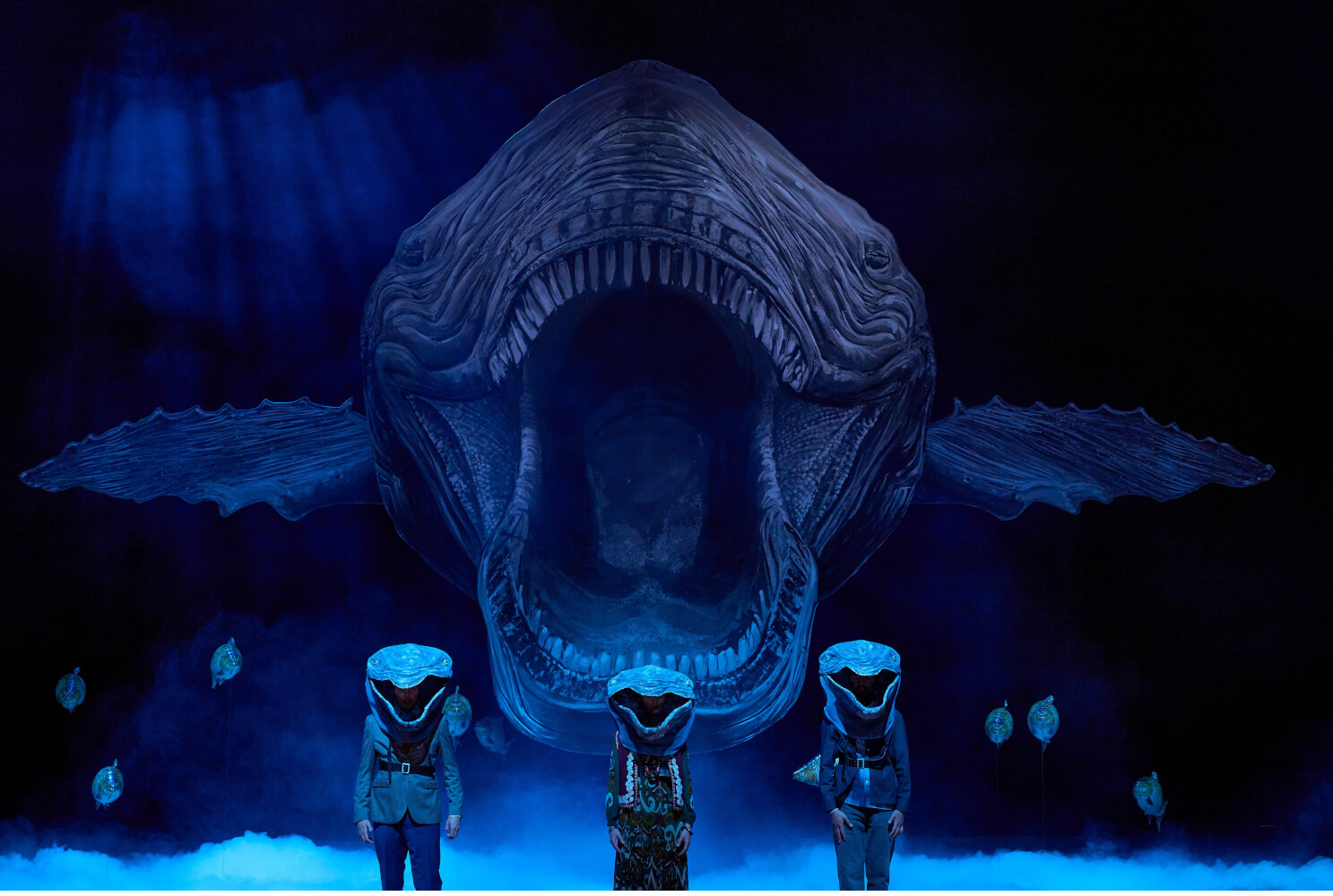 Im Hintergrund reist ein riesiger Wal bedrohlich sein Maul auf. Davor stehen drei Spieler:innen, die ebenfalls Walköpfe tragen.