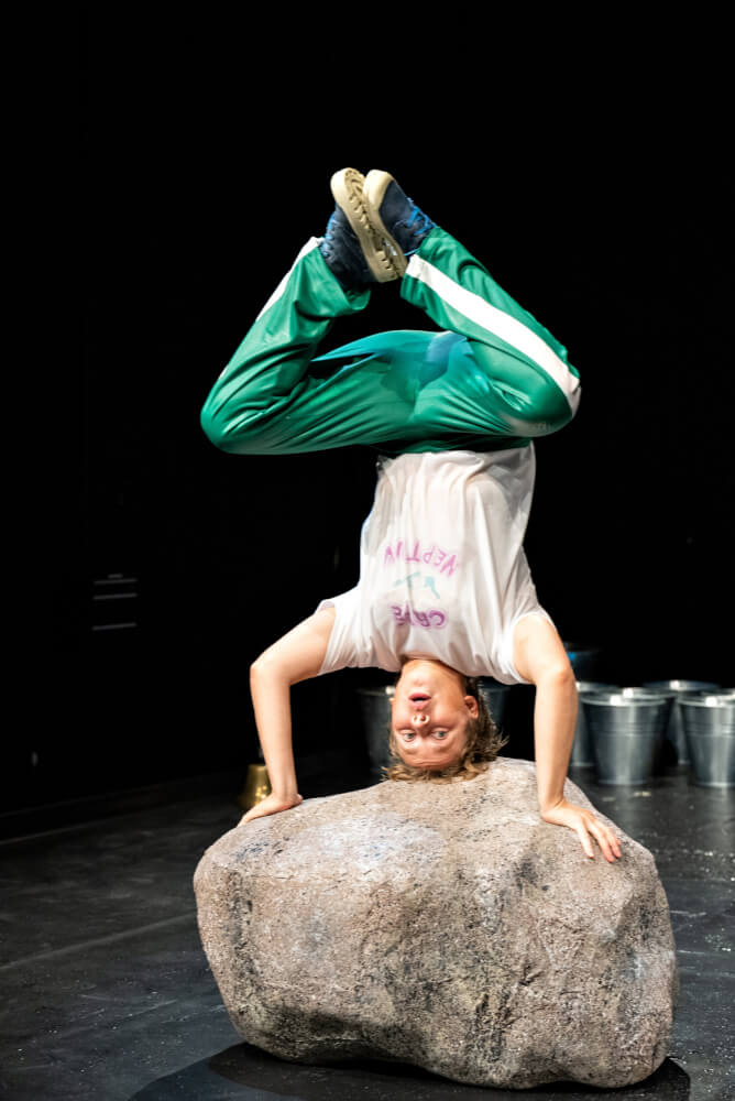 Zu sehen ist eine Schauspielerin, die auf einem massiven Stein einen Kopfstand macht.