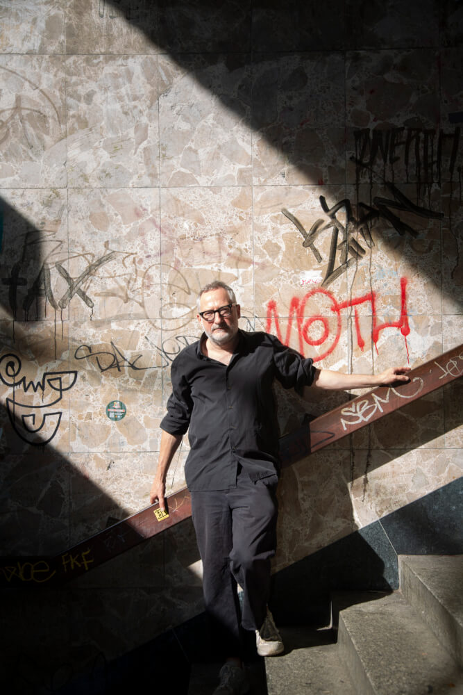 Zu sehen ist der Autor Roland Schimmelpfennig vor einer glatten Steinplattenwand, auf einer Treppe stehend und sich mit den Händen am Handlauf festhaltend.