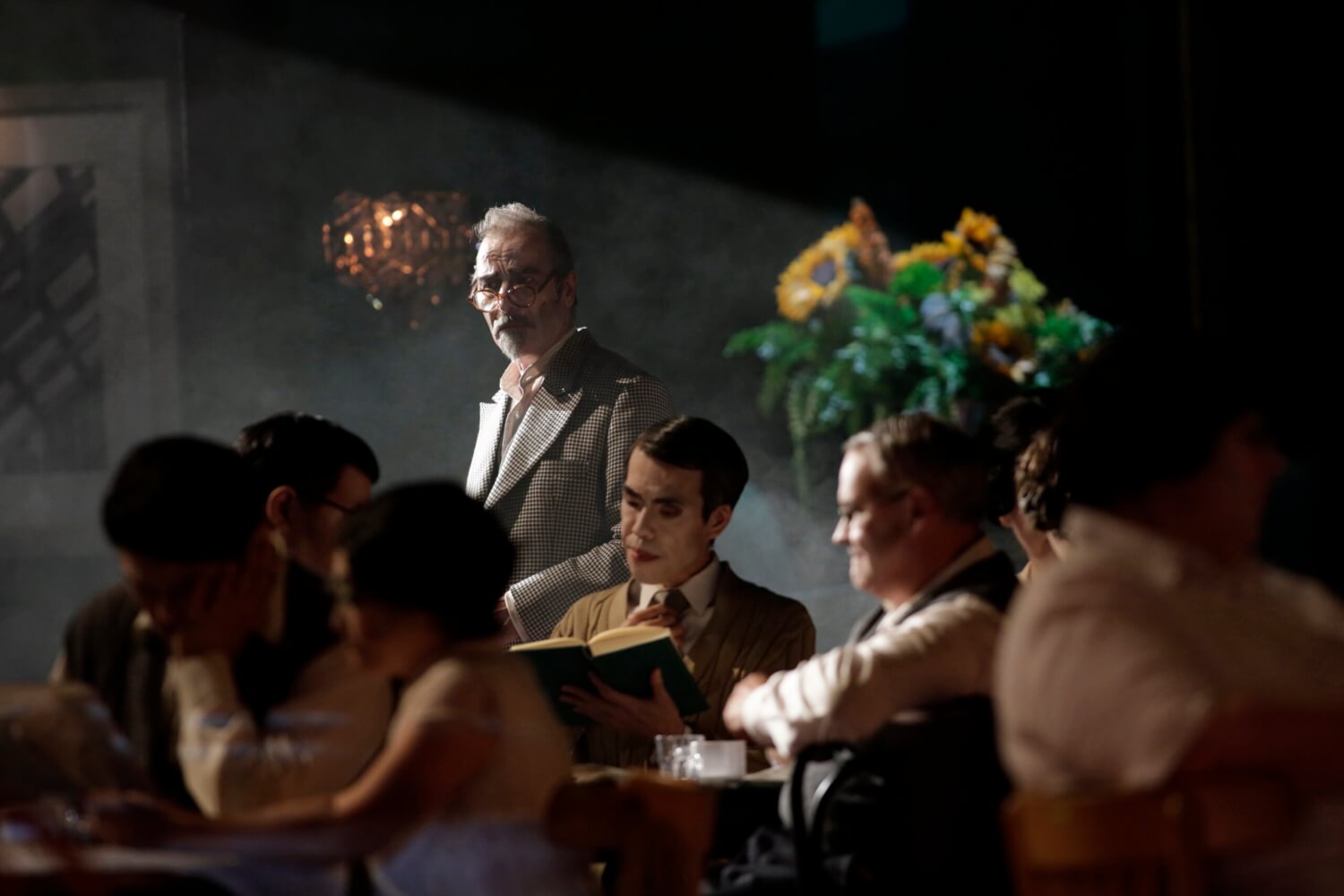 Im Vordergrund sitzt der Opernchor und unterhält sich angeregt. Im Hintergrund steht Henry Meyer und blickt über das Geschehen.