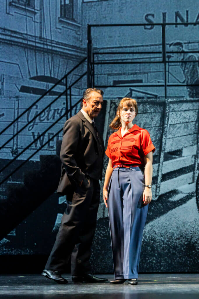 Szenenfoto aus „Das Glück ist eine Orange“ am Landestheater Detmold von Thomas Zaufke und Peter Lund. Ein Mann im dunklen Anzug geht hinter einer Frau entlang und beäugt sie dabei. Sie trägt eine rote kurzärmelige Bluse und eine blaue Stoffhose und wendet sich skeptisch von ihm ab.