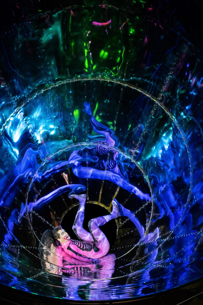 Weltuntergangs-Musiktheater „Le Grand Macabre” von György Ligeti am Staatstheater Wiesbaden. Ein Mann in einem rosa-lila-farbenen Ganzkörperanzug liegt in einem silberglänzenden Spiraltunnel.