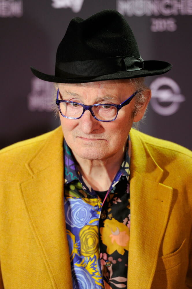 Herbert Achternbusch mit schwarzem Hut und ockerfarbenem Jackett. Darunter ein bunt geblümtes Hemd.