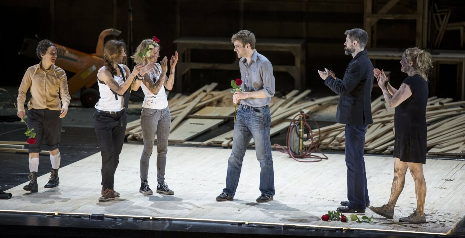 Schlussapplaus von „Die lächerliche Finsternis“ auf den Mülheimer Theatertagen. Drei Schauspielerinnen links und ein Mann und eine Schauspielerin rechts applaudieren Wolfram Lotz in der Mitte, der eine rote Rose in den Händen hält.