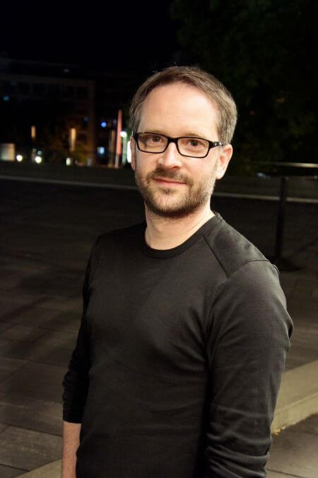 Zu sehen ist ein Porträt von Benjamin Künzel mit schwarzer Rahmenbrille und schwarzem Oberteil.