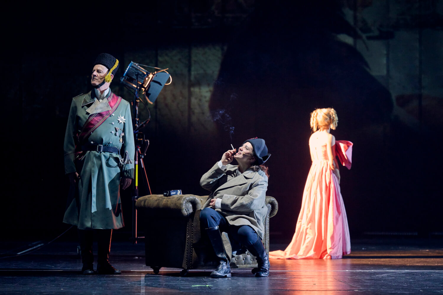 Aufführungsfoto von „Napoleon“ von Armin Petras am Saarländischen Staatstheater. Ein weiblicher Napoleon sitzt auf einem Sessel und raucht. Links daneben steht ein Mann in Uniform. Im Hintergrund steht eine blonde Frau im apricotfarbenem Kleid mit dem Rücken zu uns.