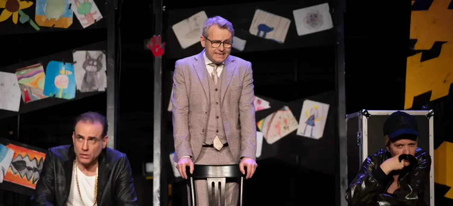 Drei Schauspielende auf einer Bühne, der mittlere stehend mit den Armen auf einem Stuhl abgestützt, der vor ihm steht