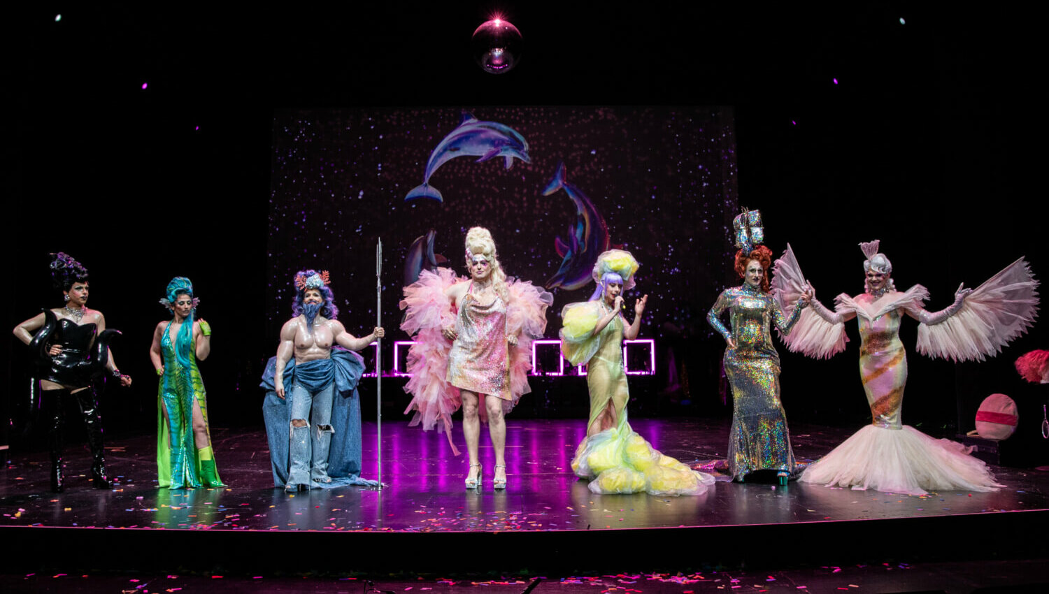 Das Drag Ensemble steht in bunten Kostümen aufgereiht auf der Bühne.