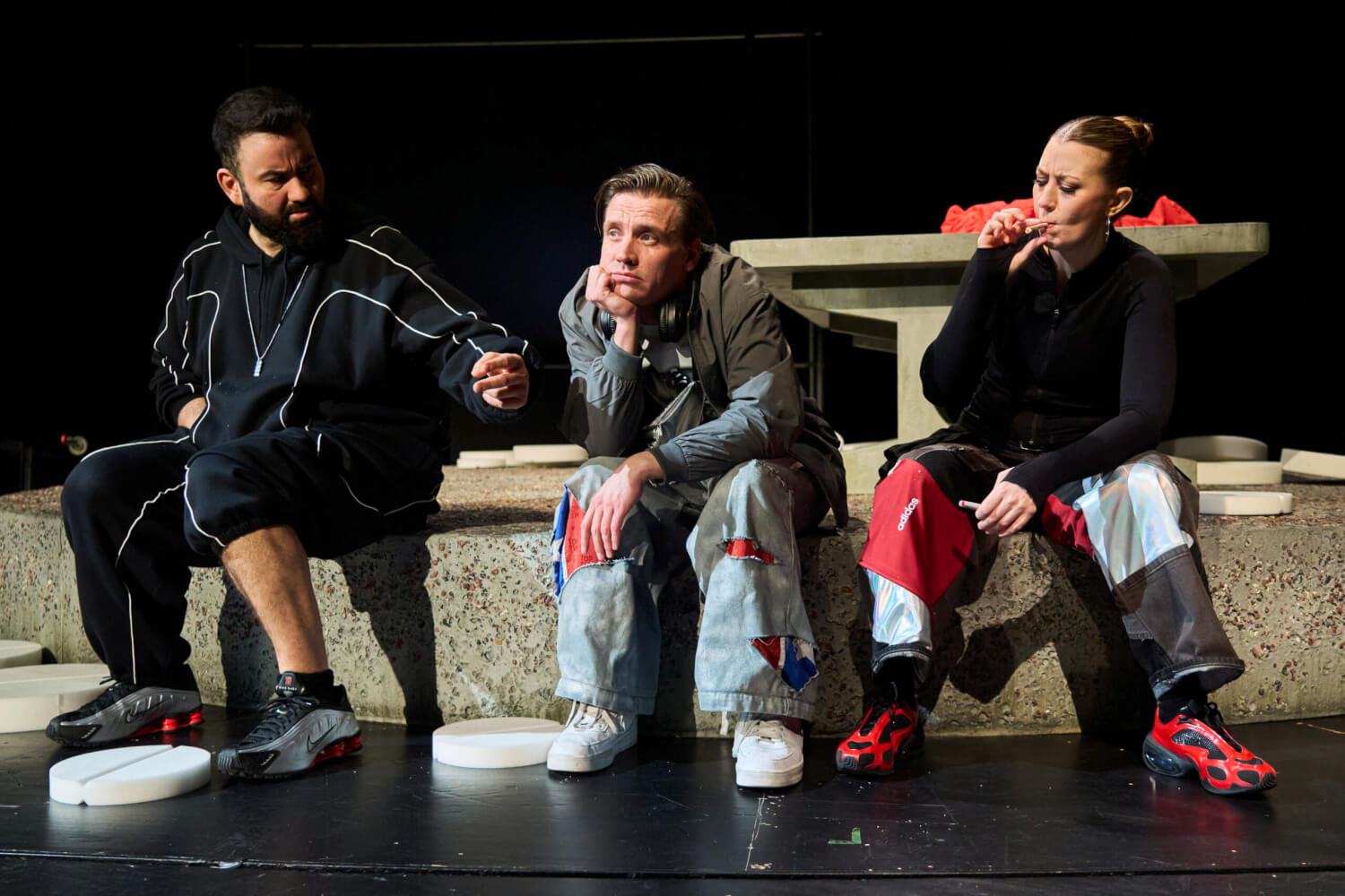 Aufführungsfoto von „Keine Aufstiegsgeschichte“ Stückentwicklung von Marco Damghani, Olivier David und Ensemble am Ernst Deutsch Theater. Zwei Männer und eine Frau sitzen auf einem Betonklotz. Die Frau raucht eine Zigarette.