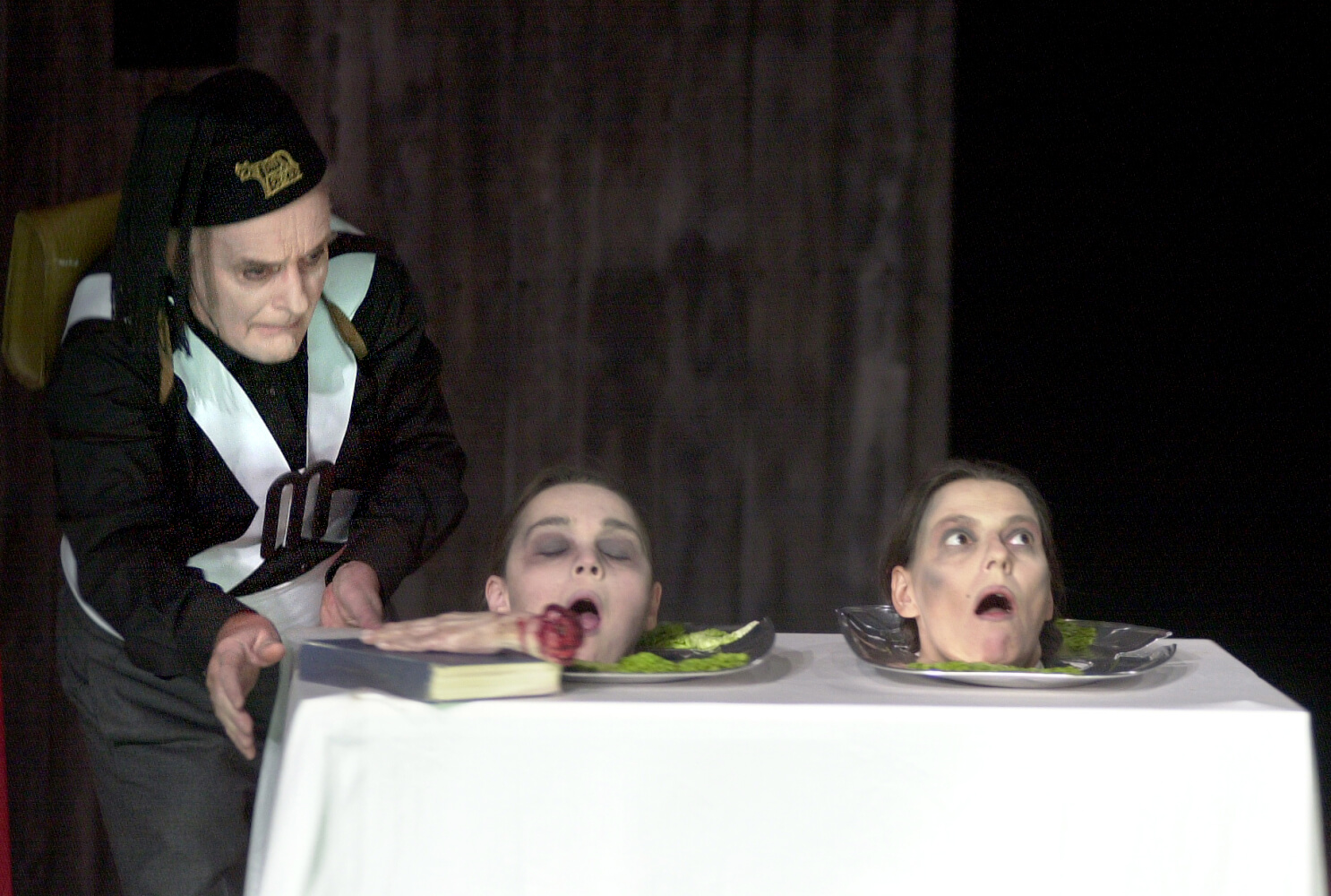 Aufführungsfoto von „Titus Andronicus“. Ein Mann steht rechts neben einem Tisch, auf dem zwei Köpfe auf Tellerplatten liegen, beide mit geöffneten Mündern.
