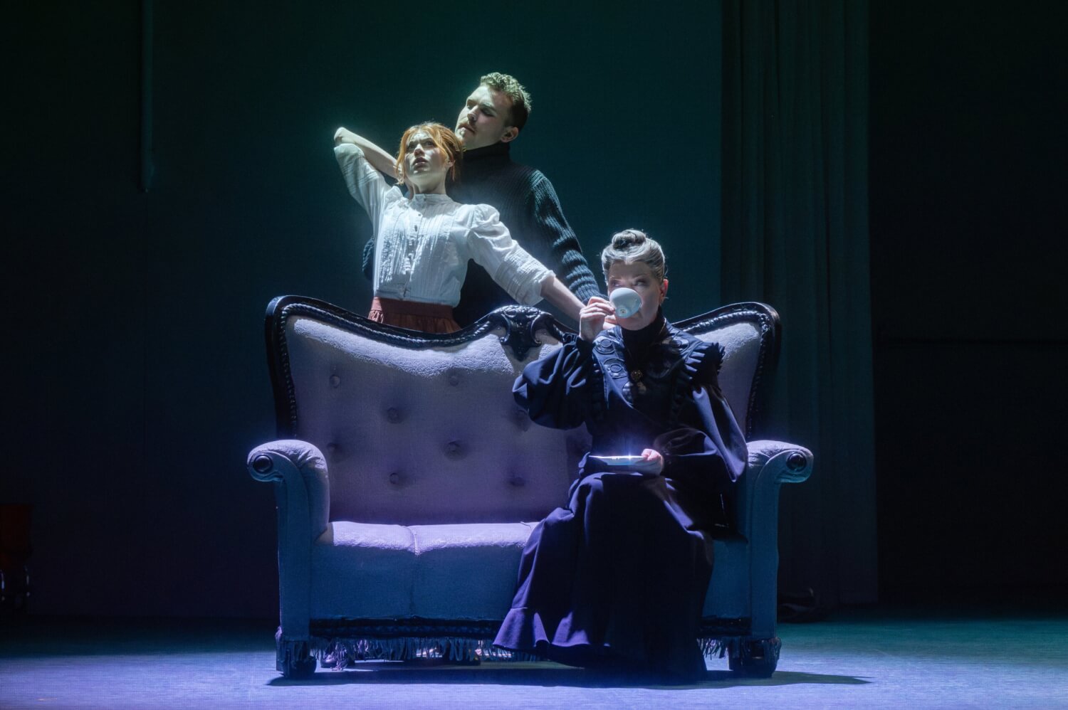 Aufführungsfoto von „Coming Up for Air“ von Bernd Franke an der Oper Leipzig. Eine Frau sitzt Tee trinkend auf einem alten Sofa. Hinter dem Sofa hält ein Mann eine Frau fest.