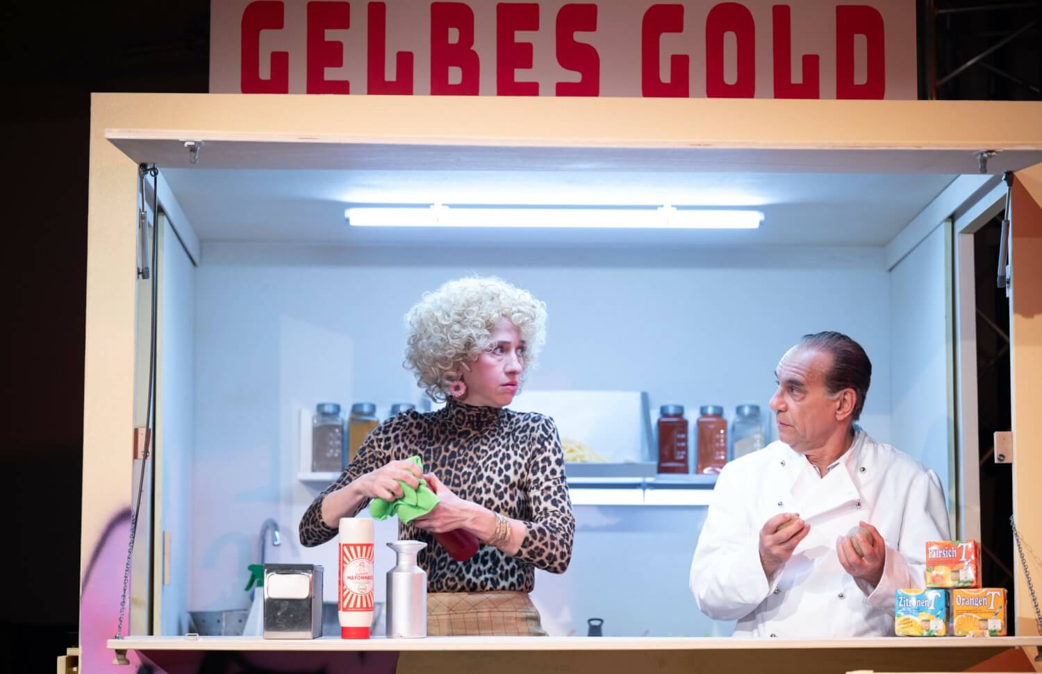 Aufführungsfoto von „Gelbes Gold“ von Fabienne Dür am Schauspiel Stuttgart. Eine Frau und ein Mann stehen in einem Imbissbüdchen mit der Aufschrift "Gelbes Gold".