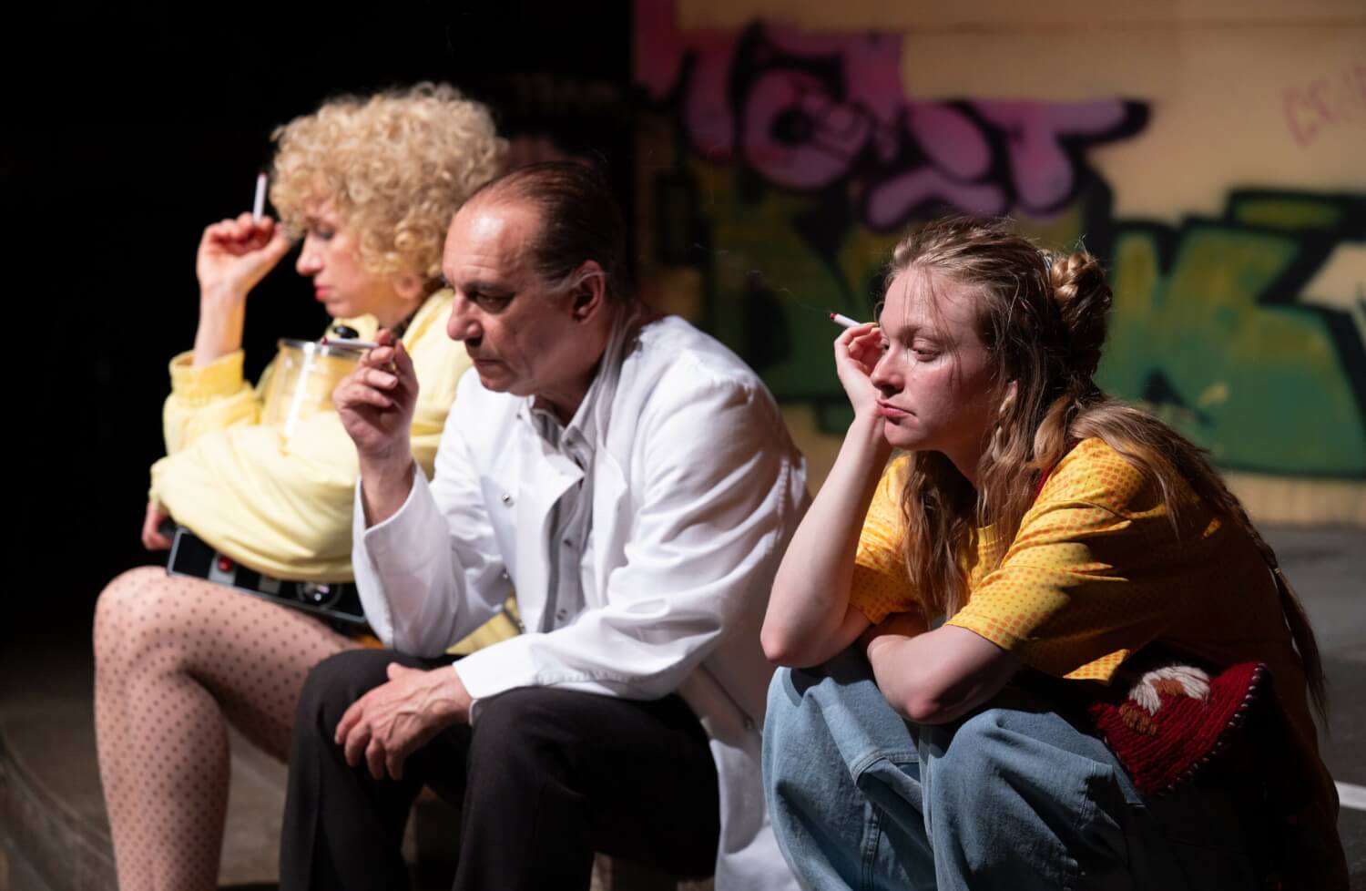 Aufführungsfoto von „Gelbes Gold“ von Fabienne Dür am Schauspiel Stuttgart. Eine Frau, ein Mann und eine junge Frau sitzen nebeneinander, alle drei rauchen eine Zigarette und wirken sehr deprimiert.