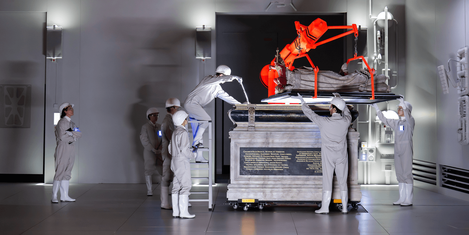 Aufführungsfoto von „Of One Blood“ von Brett Dean an der Bayerischen Staatsoper. Mehrere Menschen in hellen Schutzanzügen und weißen Helmen bauen an einem Sarkopharg.