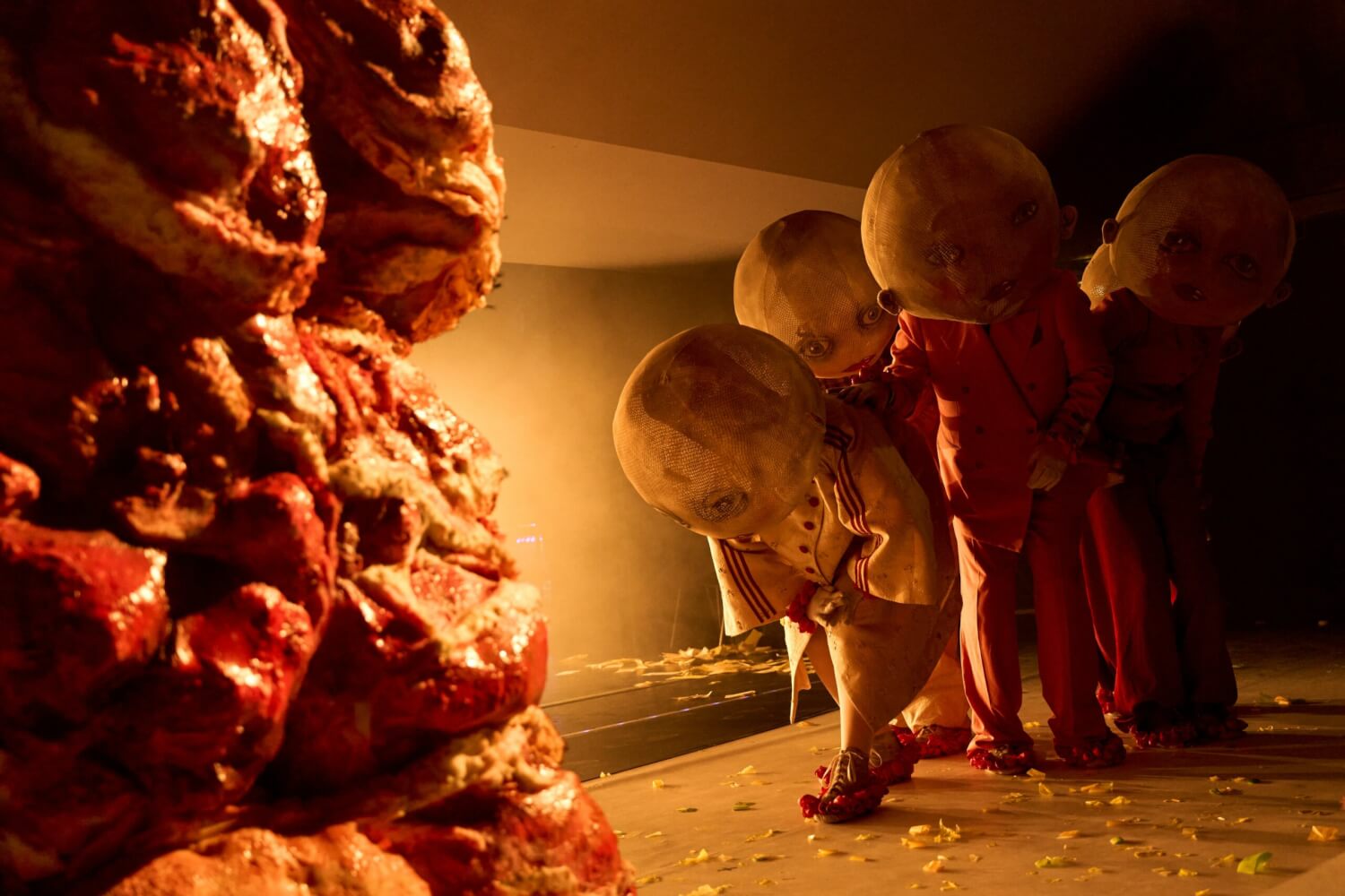 Vorne links steht ein riesiger Berg Fleisch. Hinten rechts steht das Ensemble mit übergroßen kugelförmigen Masken in eine Gruppe gedrängt.