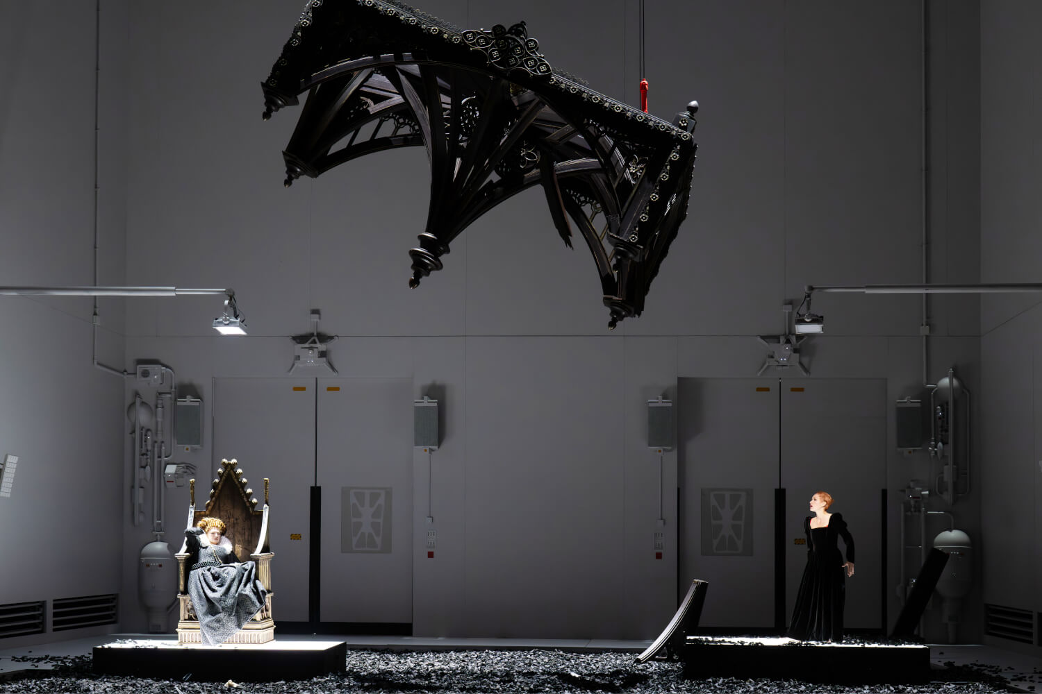 Aufführungsfoto von „Of One Blood“ von Brett Dean an der Bayerischen Staatsoper. Eine Frau links sitzt auf einem Thron, rechts steht eine Frau und schaut zu ihr. Über ihnen schwebt ein riesiges Konstrukt einer Fassade.