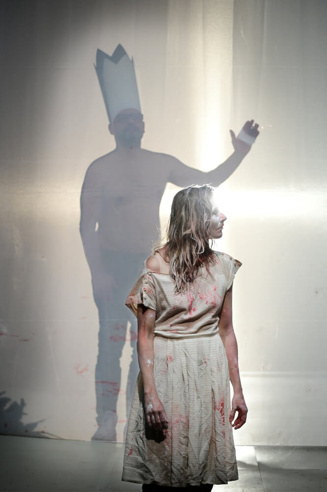 Julischka Eichel in „come as you are“. Eine Frau steht in einem weißen, rot beschmiertem Kleid und schaut nach rechts. Hinter ihr steht ein Mann, oberkörperfrei mit einer hohen, weißen Krone auf dem Kopf, der die linke Hand hebt.