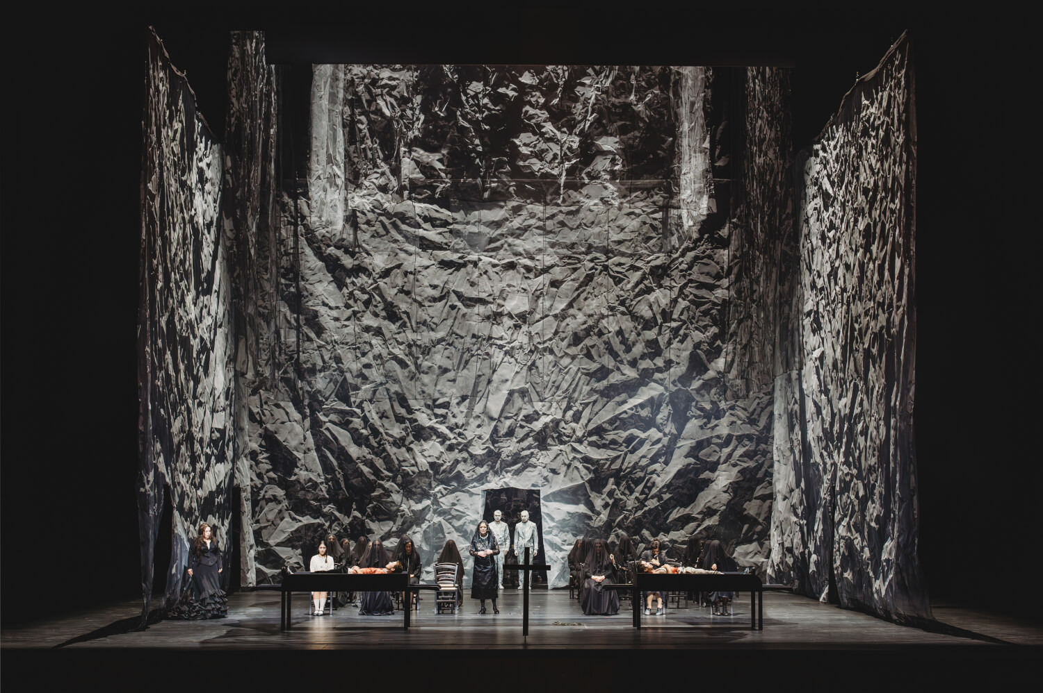 Aufführungsfoto von „Bluthochzeit“ von Wolfgang Fortner an der Oper Frankfurt. Ein sehr hoher Bühnenraum, der mit grauem, zerknitterten Stoff behängt ist, der aussieht wie Felswände. Am Boden zwei Tische au denen jeweils ein Mensch liegt, ein Kreuz in der Mitte, mehrere Menschen stehend.