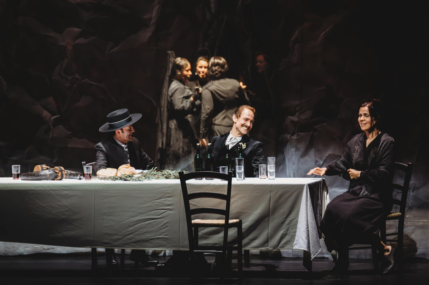 Aufführungsfoto von „Bluthochzeit“ von Wolfgang Fortner an der Oper Frankfurt. An einer langen Tafel mit weißem Tischtuch sitzen zwei Männer nebeneinander. Am Kopf des Tisches sitzt eine Frau.