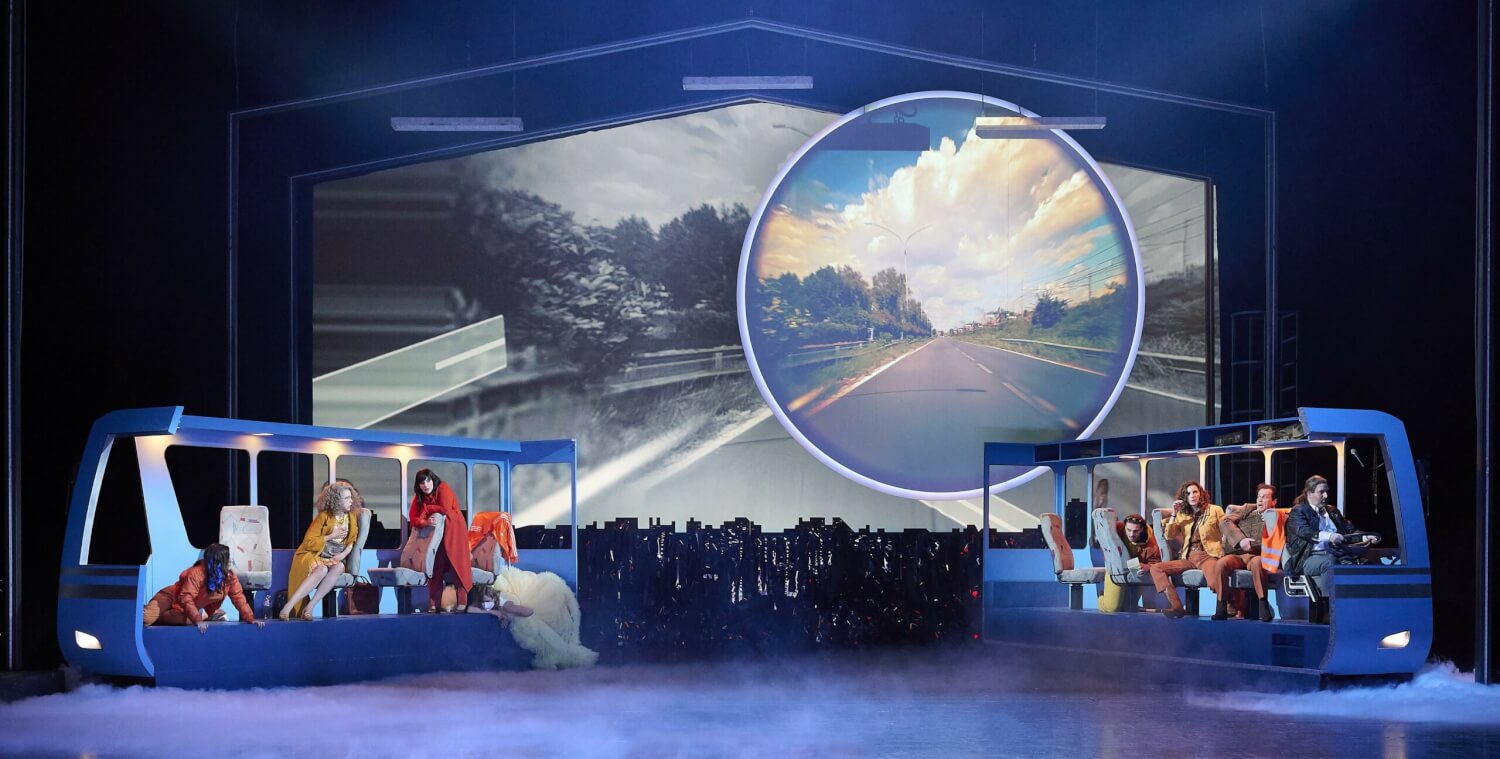 Aufführungsfoto von „Station Paradiso“ von Sara Glojnarić an der Staatsoper Stuttgart. Im Hintergrund eine Leinwand mit einer Straße. Im Vordergrund ein schematisch dargestellter Bus, der in der Mitte geteilt ist und dessen zwei Hälften links und rechts platziert sind. Darin sitzen der Busfahrer und Fahrgäste.