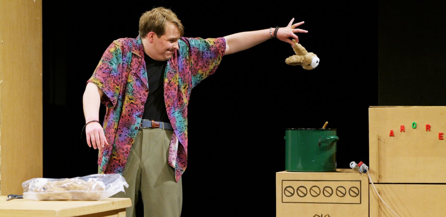 Aufführungsfoto von „Bezahlt wird NICHT!“ von Dario Fo und Franca Rame am Harztheater. Ein Mann mit buntem Hemd hält mit gespreizten Fingern einen Kopf eines Plüschhasens fest und guckt dabei verkniffen. Er steht in einer gemalten Küche mit Tisch, Kühlschrank unf Herd, auf dem ein großer, grüner Kochtopf steht.
