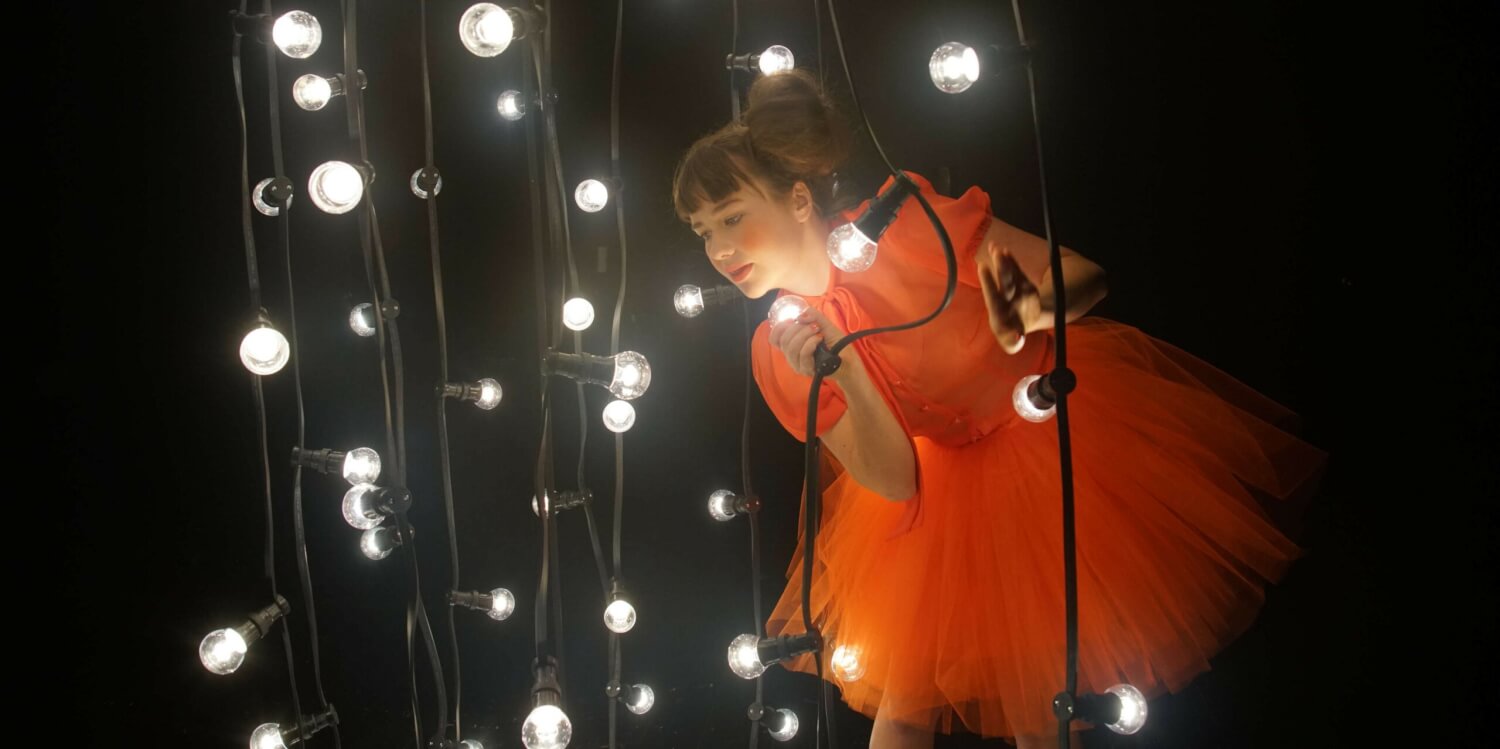 „Bilder deiner großen Liebe“ vom Landestheater Schwaben mit Franziska Roth. Eine junge Frau in einem orangefarbenen Tüllkleid steht in einem dunklen Raum, um sie herum hängen Glühlampen herunter.