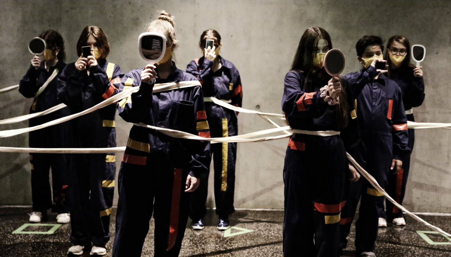 Ein performativer Spaziergang beim Festival Bohei. Sieben junge Frauen in sportlichen Anzügen stehen versetzt zueinander, mit einem hellen Band miteinander verbunden, halten sich einen Handspiegel oder ein Handy vors Gesicht.